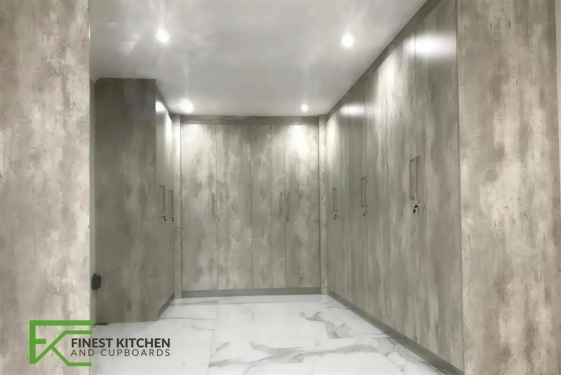 An FKC built-in cupboard run in white, set against a concrete-look feature wall above a marble floor.