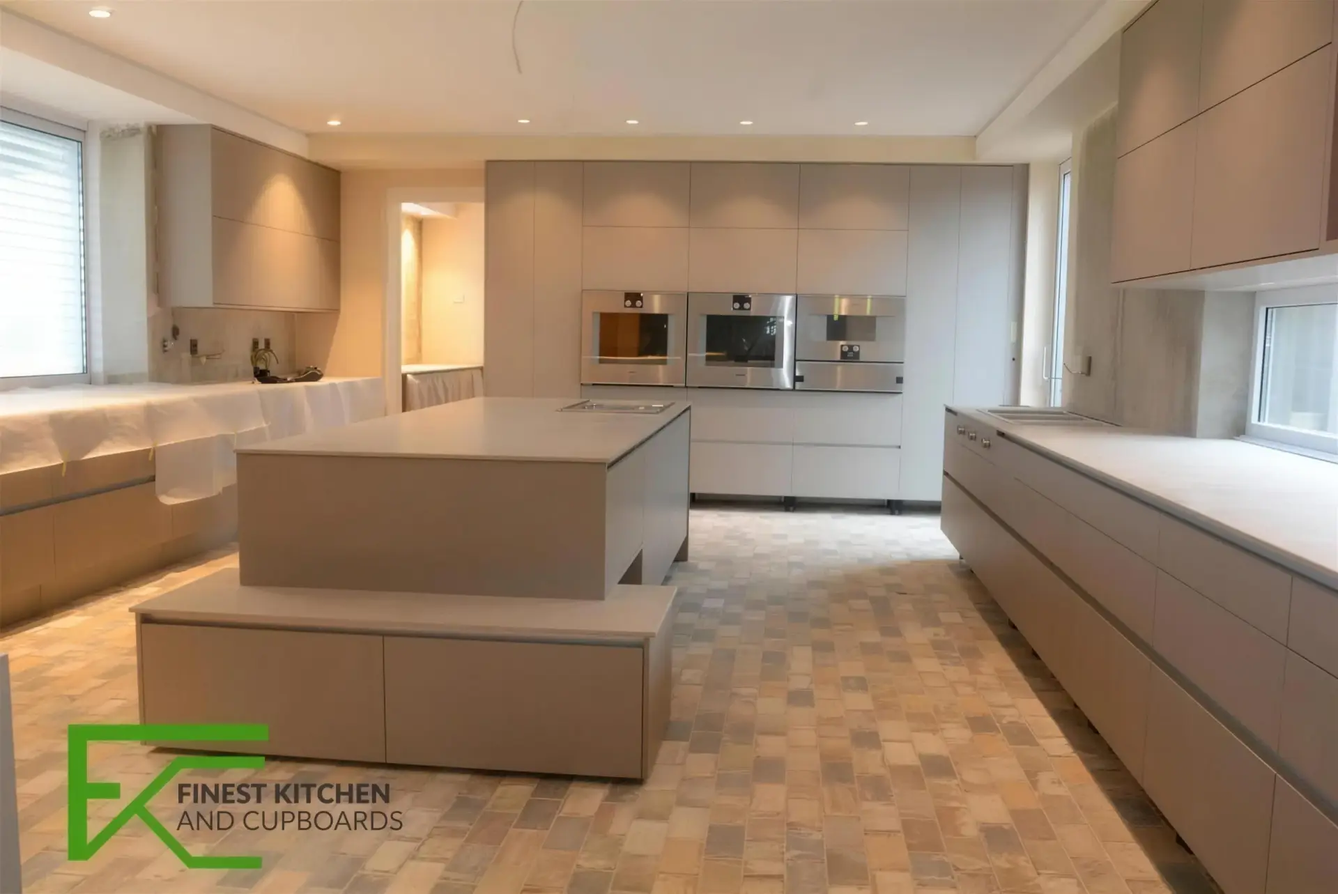 A bright FKC kitchen with a large white island, taupe base units and a wood-look tiled floor running into the living area.