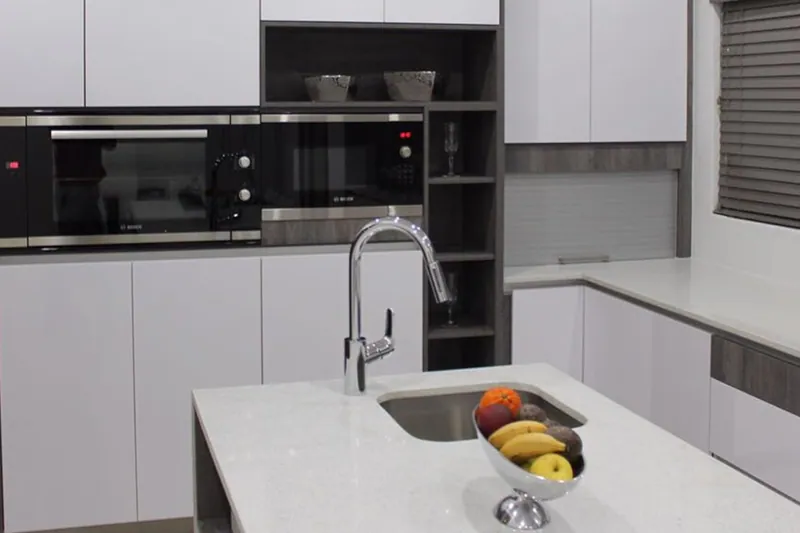 A white gloss kitchen with a black oven stack, dark timber shelving and an island with seating, built by FKC Kitchens.