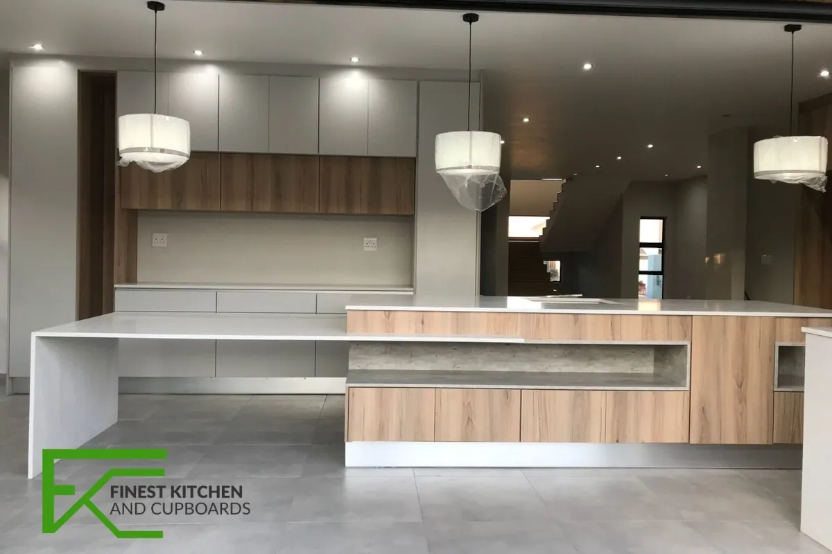 An open-plan FKC kitchen with a wood-fronted island, white stone worktop, drum pendant lights and full-height white wall units.