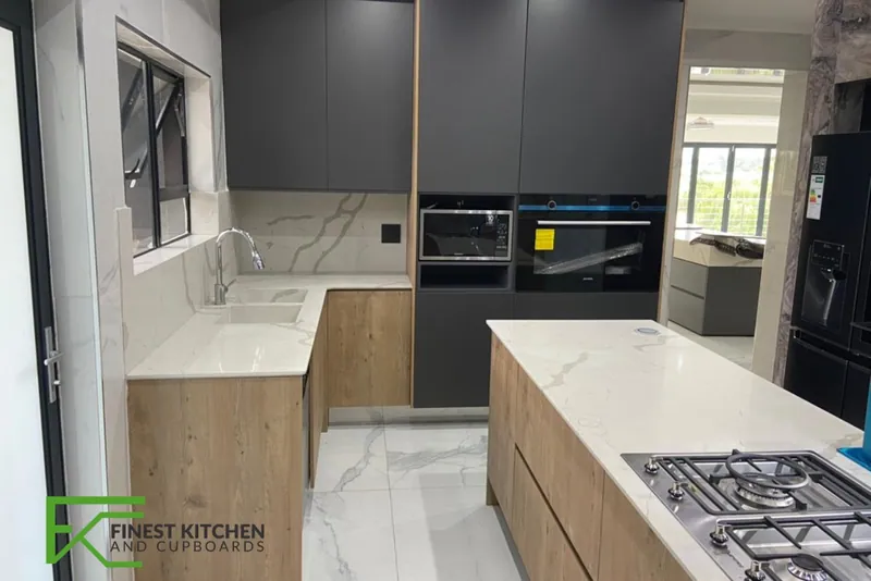 An FKC kitchen with a marble island housing a gas hob, timber and dark grey base units and a black oven stack.